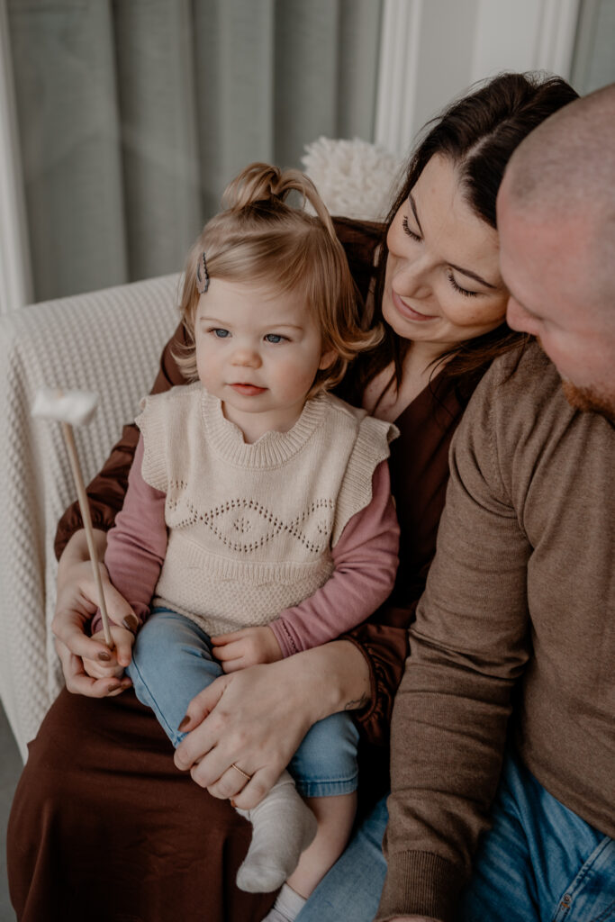Gezin dat samen met hun peuter geniet van een warme winter minisessie bij Loodz Mosa in Beers, terwijl het meisje nieuwsgierig naar een marshmallow op een stokje kijkt.