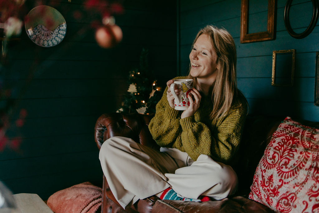 Vrouw die ontspannen met een warme mok op de bank zit tijdens een knusse kerstshoot in een huisje in Boekel, met zachte lichtjes en een warme wintersfeer, als onderdeel van haar branding.