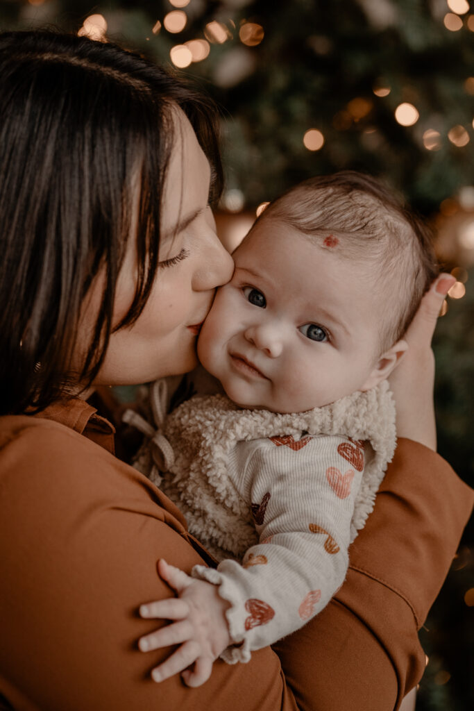 Kerst minishoot Beers: moeder houdt haar baby vast en geeft een kus voor de kerstboom tijdens een familieshoot bij Loodz Mosa.