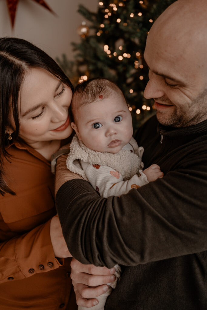 Ouders die hun baby dicht vasthouden tijdens een kerst minishoot bij Loodz Mosa in Beers, met warme lichtjes van de kerstboom op de achtergrond en een zachte, intieme sfeer.