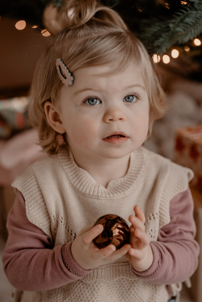 Peutermeisje dat een kerstbal vasthoudt tijdens een warme kerstshoot bij Loodz Mosa in Beers, met zachte lichtjes van de kerstboom op de achtergrond.